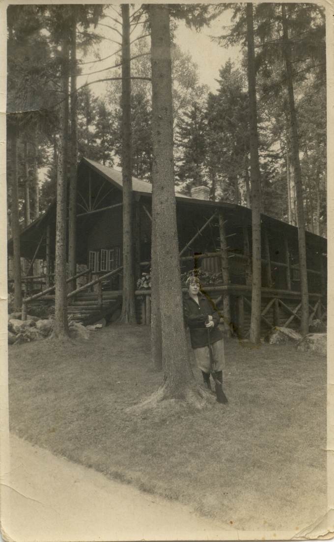 A log cabin among tall pines, with a figure at the corner